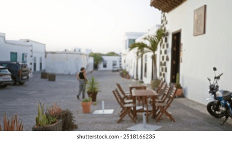 Blurred woman walking outdoors in lanzarote, spain with defocused background featuring white buildings, potted plants, parked cars, and outdoor seating on a paved street - Powered by Shutterstock