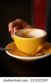 Blurred Woman's Hand Reaches For Half Empty Yellow Cup Of Coffee On The Table