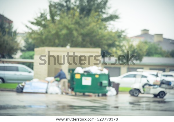 Blurred worker man collects garbage of overflowing\
dumpster at apartment building in Humble, Texas, US. Overfilled, on\
ground full of household waste, plastic bag, boxes after holidays.\
Vintage tone.
