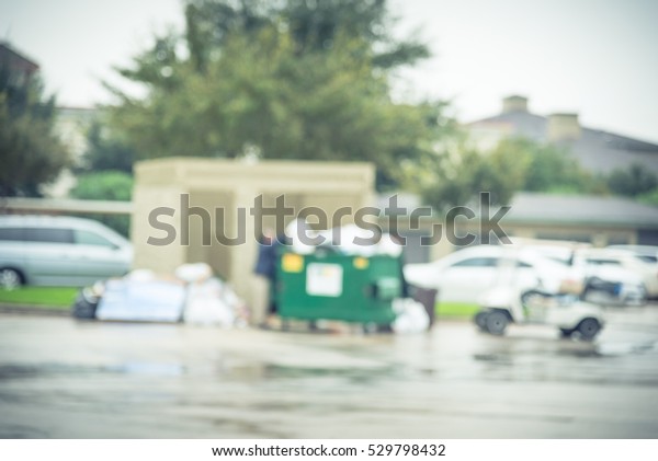 Blurred worker man collects garbage of overflowing\
dumpster at apartment building in Humble, Texas, US. Overfilled, on\
ground full of household waste, plastic bag, boxes after holidays.\
Vintage tone.