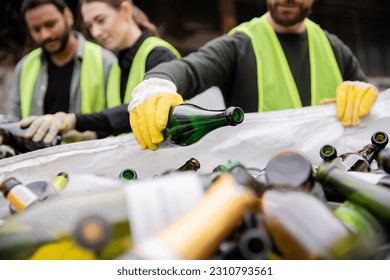 Blurred worker in protective vest and gloves putting glass bottle in sack while working with multiethnic colleagues outdoors in waste disposal station, garbage sorting and recycling concept - Powered by Shutterstock