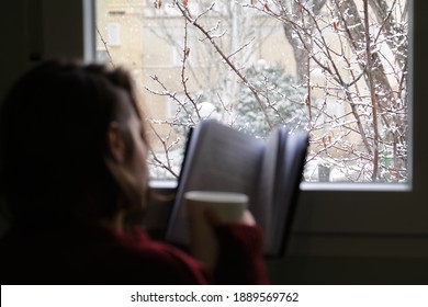 Blurred Young Woman Holding A Hot Coffee Or Tea Cup, Reading A Book By The Window In A Snowy Winter Day. Cozy At Home Concept.