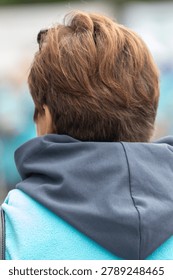 Blurry background frames a young person's back. Brown hair curls softly around the neck. Blue hoodie contrasts with gray hood lining. Person stands still, facing away from the viewer