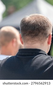 Blurry background shows two men outdoors. One man's back faces the camera. Short hair, black shirt, and earpiece visible. Sunlight highlights the scene gently. Focus on the man's head and shoulders