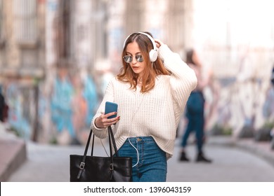 Unscharfes oder unscharfes Bild: Schönes, attraktives junges Mädchen in Jumper und Jeans mit Kopfhörer Smartphone und Sonnenbrille macht sich beim Gehen selbst