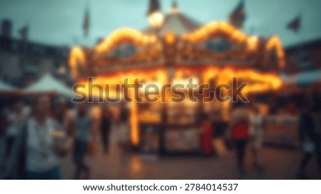 A blurry image of a carousel with bright lights at a fairground at dusk. People are walking around the carousel, enjoying the evening.