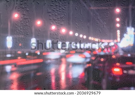 Blurry lights of moving cars and lanterns reflecting on the wet asphalt in the night city behind rain-covered window. Rainy bad autumn weather concept.