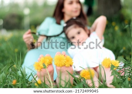 Blurry mother and daughter sitting on the grass in the park on a summer day.In the foreground are bare feet with dandelion flowers between the toes.Summer,closeness to nature,simple slow living.