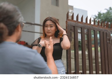 A blurry old guy in the photo is talking and pointing the young lady who's standing beside the gate, one hand on her forehead, and the other on her chest. A rail, and building in the background. - Shutterstock ID 2326116857