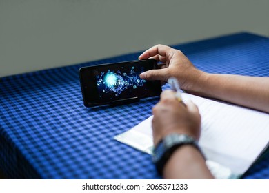 A blurry photo of a man's hand writing in front of a mobile phone showing an NFT (Non Fungible Token) image on the screen