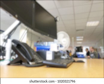 Blurry Picture Of An IT Office Workspace  With Two People Sitting Working On Their Computers. There Are Also Some Monitors, Telephone, Keyboard And Electric Desk Fan All Blurred