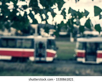 Blurry picture of two red tram carriages in Brno city with blurry leaves and intense sunlight in the top. Intentionally blurry transportation vehicles behind blurry green-white foreground elements.