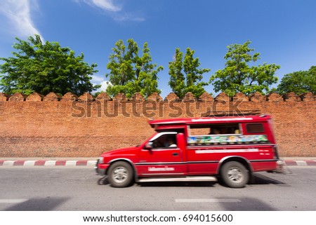 Blurry Red Car Public Taxi Running on The Road and Passing Big Brick City Wall of Chiangmai, Thailand with Blue Sky.