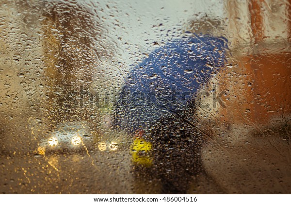A\
blurry silhouette with umbrella on a city street seen through wet\
window with focus on some water drops. Toned\
effect
