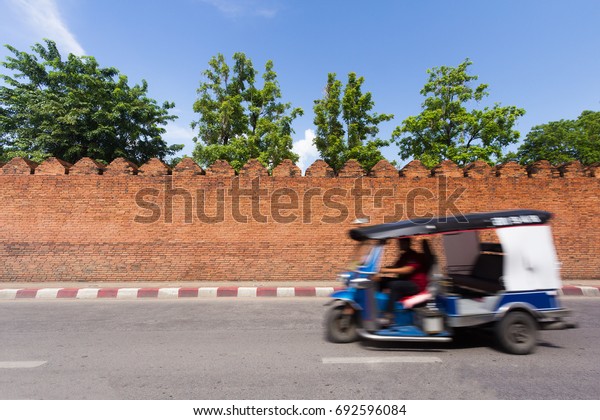 Blurry Three Wheels Taxi (TUK TUK) Running on\
The Road and Passing Big Brick City Wall of Chiangmai, Thailand\
with Blue Sky. Copy Space for\
Text.