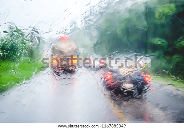 Blurry\
vehicle silhouette seen through car window with rain drops on the\
road cut through the mountains and green trees along the way, cars\
are overtaking heavy trucks while rain is\
falling