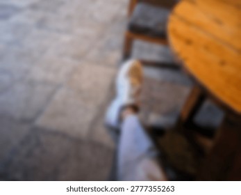 A blurry view looking down at a casual white sneaker worn by a person sitting at a small wooden table on a stone patio cafe terrace.