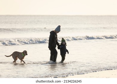 Blyth, UK - Dec. 10, 2021: A Mother And Son Enjoy The Water And The Beach With Their Dog, Protected By Cold Weather Clothing And Boots, North Sea, Northumberland