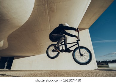 Bmx Rider Making A Bunny Hop. He Is Wearing Black Clothes And White Helmet. Black Bmx Bike. He Is Under A Modern Structure. During The Sunset.