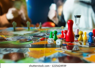 Board Game Concept- Board Game Field, Many Figures, Meeple, Dice, Coins And Sandglass. Two People Play Holding Cards On Blurred Background