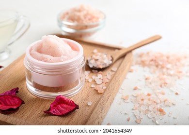 Board with jar of body scrub, sea salt and flower petals on light background