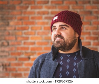 Boarded guy in a woolen hat and sweater smiling on a brick wall.