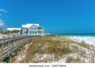 Boardwalk and beach house near the white sand shore of a beach at Destin, Florida. There are grasses on the white sand and a view of a gray house with terrace at the back. - Shutterstock ID 2205730415