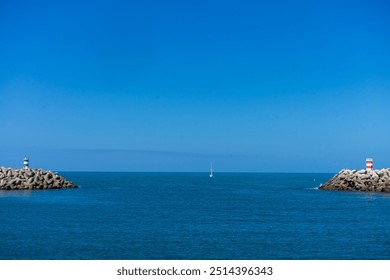 A boat approaches the entrance of Nazaré Marina, Portugal, with the lighthouse towers on both piers clearly visible, against a backdrop of calm waters and clear skies. - Powered by Shutterstock