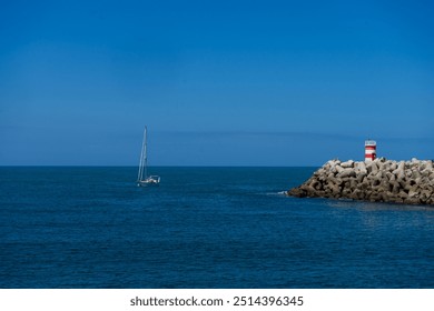 A boat approaches the entrance of Nazaré Marina, Portugal, with the lighthouse towers on both piers clearly visible, against a backdrop of calm waters and clear skies. - Powered by Shutterstock