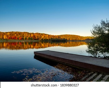 A Boat Dock In The Foreground At Keystone Lake In West Moreland Country, Nestled In The Laurel Highlands Of Pennsylvania In The Fall With The Colorful Trees Reflecting In The Lake And A Nice Blue Sky.