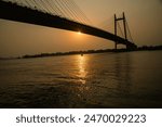 a boat floating on the water of river Ganga in the vibrant golden light during sunset beneath the largest hanging bridge of Asia calle d the Second Hoogly Bridge 