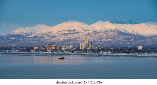 A boat in front of the Anchorage Alaska skyline with snow covered mountains in the background at sunset during winter