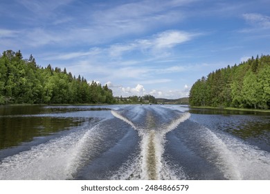 A boat leaves a V-shaped wake as it travels down a calm river bordered by lush green trees under a clear blue sky. - Powered by Shutterstock