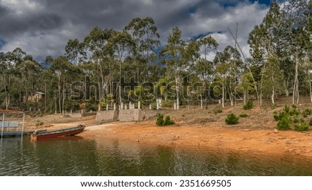  The boat is moored at the shore. Trees and bushes grow on the red soil. The flag of Madagascar is flying. Clouds in the blue sky. Mantasoa Lake. Nosy Soa Park. Lemur Island  