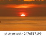 Boat sits in calm ocean seas as big orange sun sets below the horizon line.