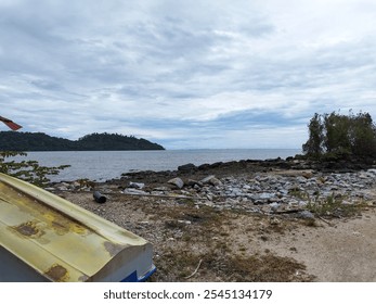 A boat sits on a rocky shore, overlooking a calm sea with distant hills. - Powered by Shutterstock