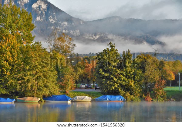 boats in a cover on the\
pier near the river and cars on the side of the road and fog in the\
mountains\

