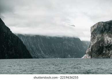 Boats Full Of Tourists Sailing Through The Water Of The Great Lake Between The Impressive Rocky Fjords Under A Sky Full Of Gray Clouds, Milford Sound, New Zealand