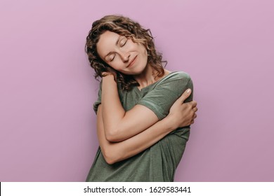 Boddy positive concept. Young curly woman hugging herself and feel calm and happy, love herself. Positive human emotions. Isolated on purple background.