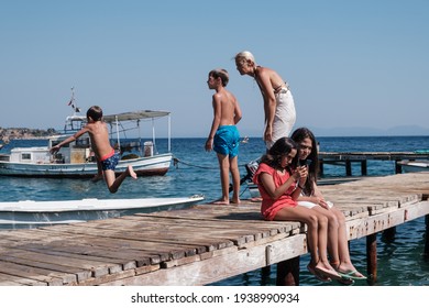 Bodrum, Turkey  - August, 2020: Kid Jumping Into The Water Leaves His Brother And Mother Behind. Two Girls Sitting On The Other Side Of The Pier.