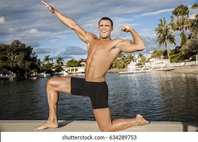 Body Builder Sitting With One Leg And Smiling, Outside Area Near Lake And Houses, Natural Trees Give Beauty To Place, Sunlight Around The Scene, Weather Is Good.