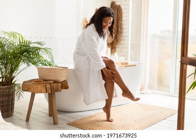 Body Care. Cheerful African American Woman In Bathrobe Touching Smooth Legs Moisturizing Skin With Aftershave Lotion Sitting On Bathtub In Modern Bathroom At Home