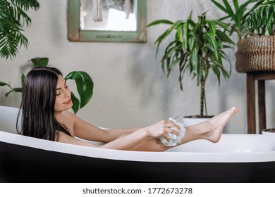 Body care concept. Side view of smiling young adult girl taking bath, using loofah and scrub legs, enjoying morning routine and spending time in bathroom - Powered by Shutterstock