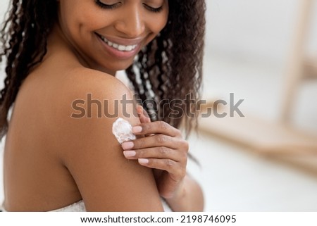 Body Care Concept. Smiling Beautiful Black Woman Applying Moisturizing Lotion On Shoulder, Happy African American Female Making Beauty Treatments While Standing Wrapped In Towel After Bath, Closeup