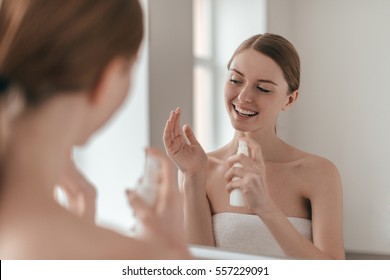 Body Care. Over The Shoulder View Of Young Beautiful Woman Applying Water Spray While Standing In Front Of The Mirror