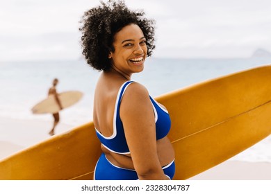 Body confident female surfer stands on the beach in a bikini, holding her surfboard and ready for an exciting adventure. Happy young woman smiling at the camera on her way to ride with the waves. - Powered by Shutterstock