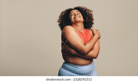 Body confident woman embracing her body, expressing self-love and self-acceptance. Young plus-size woman standing in a studio in fitness clothing, embracing her natural physique with joy.
