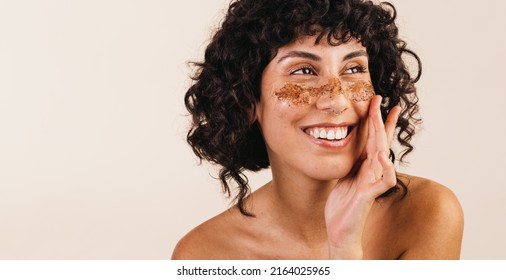 Body Conscious Young Woman Looking Away With A Smile While Applying Coffee Scrub On Her Face. Happy Young Woman Exfoliating Her Facial Skin Using A Cleansing Coffee Mask.