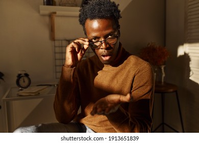 Body language. Picture of shocked stylish young dark skinned man sitting in cozy room, lowering spectacles, making questioning gesture with palm, opening mouth in indignation and outrage