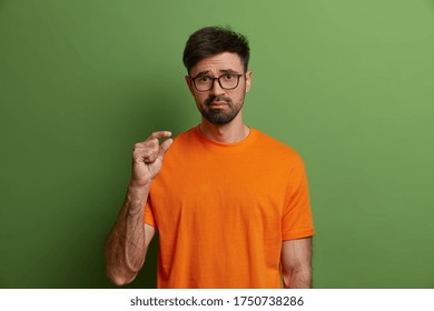 Body Language And Size Concept. Young Displeased Man Demonstrates Tiny Measure, Tells About His Little Salary, Shapes Something Small, Judges Poor Quality, Wears Orange T Shirt, Isolated On Green Wall